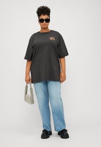 Dunkelgraues, übergroßes T-Shirt mit kurzen Ärmeln, das ein kleines orangefarbenes Graphic zeigt. Getragen mit hellblauen, stark verwaschenen Schlagjeans und schwarzen Plateauschuhen.