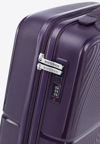 Purple hard-shell suitcase featuring a textured surface, double zipper with silver tags, and a combination lock on the side.