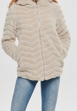 Veste beige en fausse fourrure avec motif en chevrons, col festonné, fermeture éclair avant, mains dans les poches latérales, portée avec un jean bleu.