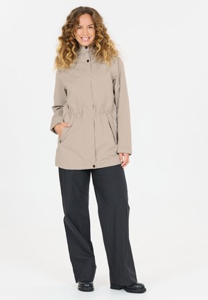 Kvinde med krøllet hår iført beige jakke med hænderne i lommerne, sorte vide bukser og sorte sko, smilende mod hvid baggrund.