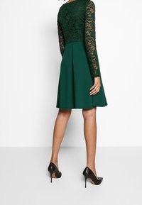 Grünes Kleid mit einem taillierten Spitzenoberteil und langen Ärmeln, ausgestelltem Rock und kniehohem Saum. Kombiniert mit schwarzen High Heels.