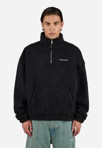 Hombre con un pulóver de forro polar negro con media cremallera y bolsillo canguro, combinado con pantalones vaqueros de color azul claro, ante un fondo liso.