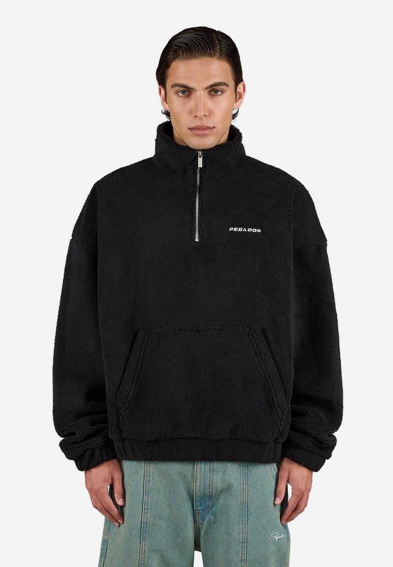 Hombre con un pulóver de forro polar negro con media cremallera y bolsillo canguro, combinado con pantalones vaqueros de color azul claro, ante un fondo liso.