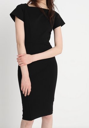 Frau trägt ein eng anliegendes, knielanges schwarzes Kleid mit kurzen Ärmeln, steht mit einem Arm über den anderen verschränkt vor einem weißen Hintergrund.