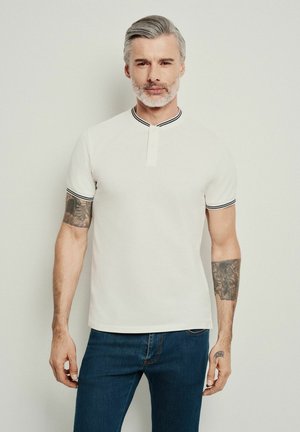 Biała koszulka polo z czarnym prążkowanym kołnierzykiem, krótkimi rękawami i zapięciem na trzy guziki. Nosi ją osoba w niebieskich dżinsach, z widocznym tatuażem na ramieniu.