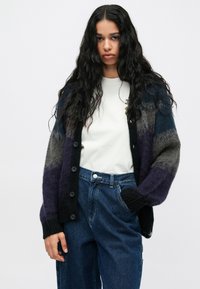 Moteris su ilgais garbanojais plaukais, apsirengusi balta marškinėliais, džinsais su aukštu liemeniu, ir mygtukais pritvirtinta pūkuota dryžuota kardiganu juoda, pilka ir purpurine spalvomis.