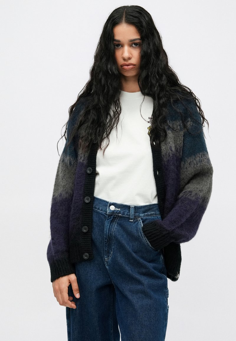 Woman with long curly hair wearing a white T-shirt, high-waisted blue jeans, and a buttoned, fuzzy striped cardigan with black, gray, and purple tones.