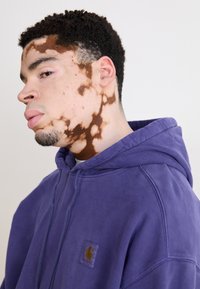 Sudadera morada con un bolsillo frontal y cordones; textura suave. Parche con el logo de la marca notable en el pecho. El modelo presenta un patrón de piel prominente.