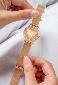 Orologio in oro rosa con quadrante triangolare, adornato con cristalli e cinturino a rete. Il quadrante presenta un design minimalista con lancette semplici.