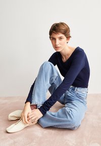 Dunkelblaues Langarmoberteil, hochgeschnittene hellblaue Jeans und weiße Schnürschuhe. Sitzpose auf einem beigen Teppich vor einem neutralen Hintergrund.