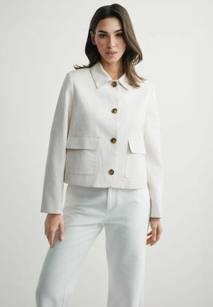 Młoda kobieta w białej, zapinanej na guziki kurtce z dużymi kieszeniami i białych spodniach, stojąca na tle jednolitego, jasnego tła.