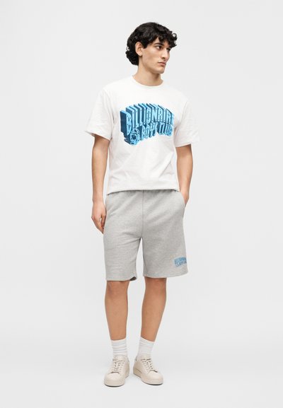 Λευκό βαμβακερό t-shirt με μπλε γραφικό κείμενο, σετ με γκρι βαμβακερά σορτς με αντίστοιχο λογότυπο, φορεμένα με ανοιχτόχρωμα αθλητικά παπούτσια.