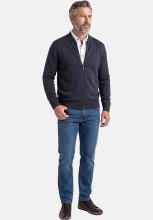 Mężczyzna z siwymi włosami i brodą, ubrany w ciemny sweter na zamek, białą koszulę z kołnierzykiem, niebieskie dżinsy i ciemnobrązowe skórzane buty, stoi na tle białego tła.