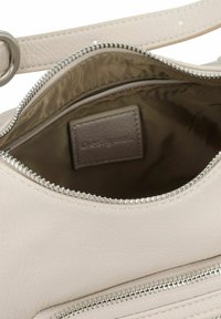Interior de un bolso de cuero beige con detalles de cremallera plateada. Incluye una etiqueta rectangular que muestra "desigual". Textura suave, diseño minimalista.
