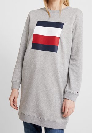 Kobieta ubrana w długą szarą bluzę z prostokątnym logo w kolorach granatowym, białym i czerwonym na piersi oraz małym logo na nadgarstku.