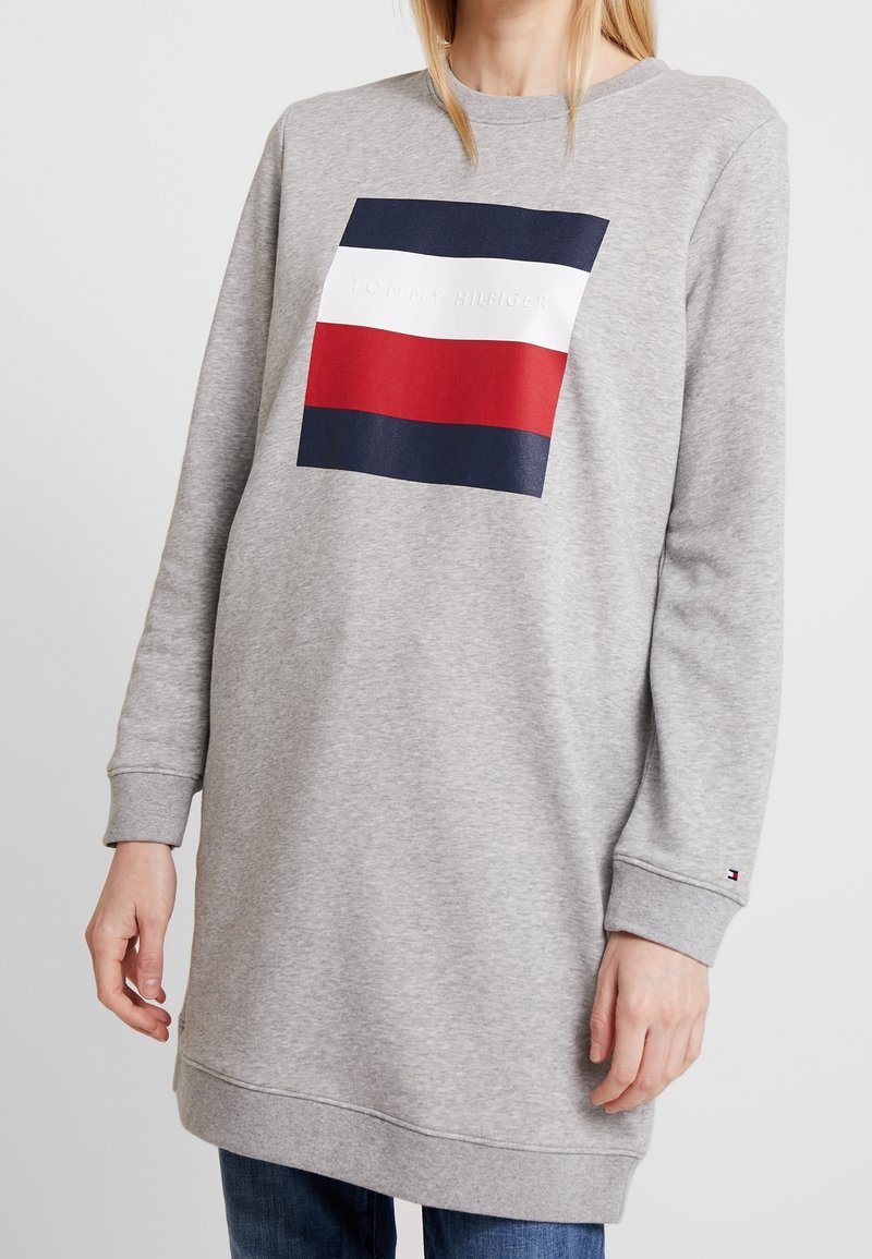 Femme portant un long sweat-shirt gris avec un logo rectangulaire bleu marine, blanc et rouge sur la poitrine et un petit logo sur le poignet.