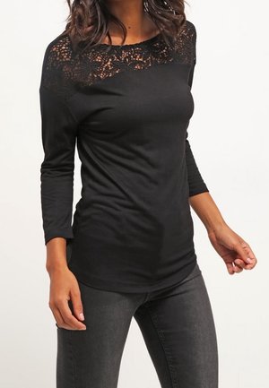 Femme portant un haut noir à manches longues avec un détail en dentelle florale sur le haut de la poitrine et les épaules, associé à un jean gris foncé.