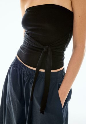 Ženska v črnem brezramnem topu s prednjim vozlom in ohlapnih temnomodrih hlačah z elastičnim pasom, z roko v žepu.