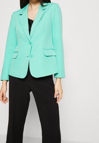 Tyrkysový blazer s přiléhavým střihem, dlouhými rukávy, zapínáním na knoflík a dvěma předními kapsami, v kombinaci s černými kalhotami.