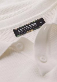 Bílá polo košile s texturou a síťovaným designem, se černou nálepkou s nápisem "ombre QUINTESSENCE" a jedním bílým knoflíkem.