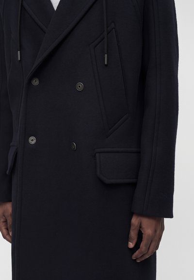 Gros plan d'une personne portant un manteau noir à double boutonnage avec des boutons métalliques et des poches à rabat, une main visible sur le côté.