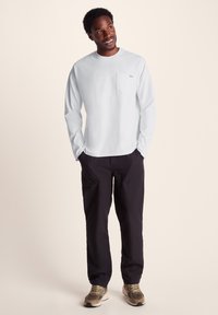 Light gray long-sleeve shirt featuring a chest pocket, paired with dark pants and neutral-colored sneakers, all against a neutral background.