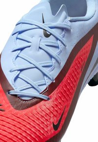 Nike jalkapallokenkä, jossa on punainen ja musta teksturoitu päällinen, vaaleansiniset nauhat, sulavalinjainen muotoilu ja erottuvat logon yksityiskohdat.
