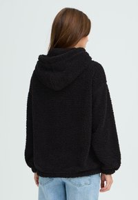 Sort fleece hættejakke med en blød, struktureret overflade, der har en hætte og en løs pasform. Synlig søm ved kanten og manchetter.