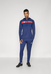 Marineblauw sport trainingspak met een rood en blauwe horizontale streep design. Inclusief een capuchon en witte zijsstrips op de broek. De stof lijkt glad.