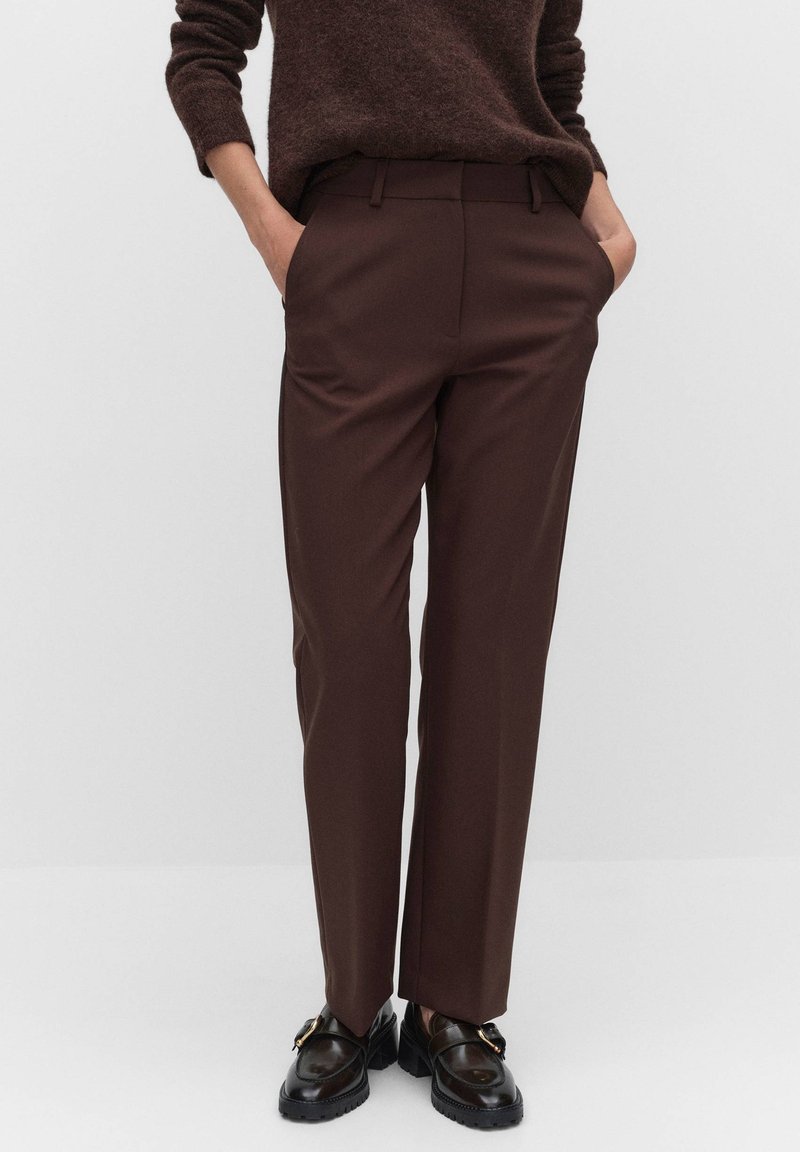 Personne portant un pantalon marron foncé sur mesure, un pull marron foncé, des mocassins noirs, les mains dans les poches, debout devant un fond clair uni.