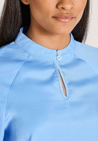 Vrouw in een lichtblauwe satijnen blouse met een keyhole halslijn bevestigd met twee kleine knopen.