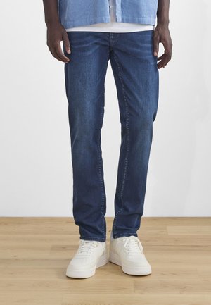 Mand iført blå denim jeans, hvide sneakers og en lyseblå skjorte, stående på et trægulv mod en hvid væg.
