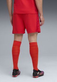 Röd idrottströja och shorts, matchande orange knästrumpor och svarta fotbollsskor med en flerfärgad design. Bakifrån.