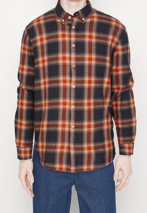 Mand iført en langærmet ternet skjorte med orange, sort og beige striber, kombineret med blå teksturerede bukser.