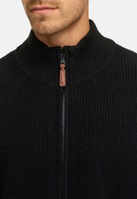 Zwarte gebreide ritstrui met een hoge kraag, voorzien van een gestructureerd patroon en een bruine leren ritsaccent.