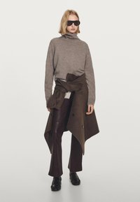 Maglione marrone a collo alto, misto lana; giacca esterna marrone annodata in vita; pantaloni in pelle marrone scuro; scarpe nere; occhiali da sole neri oversize.