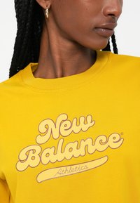 Kollane puuvillane dressipluus, millel on ümar kaelus, lilla "New Balance" logo ning lühikesed punutud juuksed koos kõrvarõngastega.