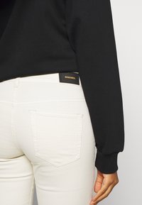 Top negro de manga larga con puños acanalados, combinado con jeans blancos que tienen un bolsillo trasero y un parche del logo negro de Diesel en la cintura.