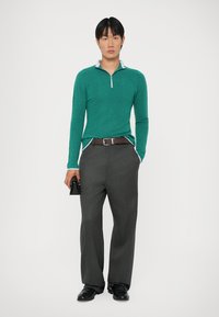 Polo verde de mangas longas com fecho éclair, colarinho azul claro; combinado com calças largas cinzas e sapatos pretos, segurando uma pequena bolsa preta.