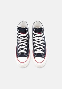 Υψηλά αθλητικά παπούτσια από ναυτικό καμβά με λευκές λαστιχένιες σόλες. Κόκκινα σημεία κατά μήκος των ραφών και του προστατευτικού μέρους των δακτύλων, με λευκά κορδόνια και λεπτομέρειες της μάρκας.