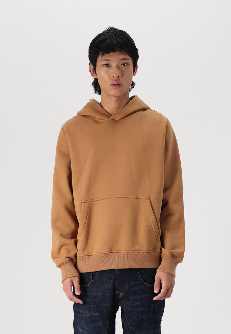 Sudadera marrón de tejido suave, con capucha y bolsillo frontal, que presenta puños y dobladillo de canalé. Combinada con jeans de mezclilla oscuros.