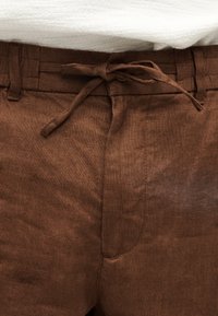 Pantalon en lin marron avec une ceinture en tissu nouée à la taille, présenté sur un fond de tissu blanc texturé.