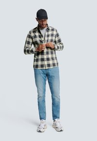 Karierte Flanellhemd in Creme, Dunkelblau und Grau; hellblaue Jeans; schwarze Mütze; schwarz-weiße Sneakers; lässiges Outfit mit Knopfdetails.