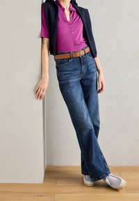 Persoon leunend tegen de muur, gekleed in een paarse shirt, zwarte trui om de schouders, blauwe jeans met een bruine riem en witte schoenen.