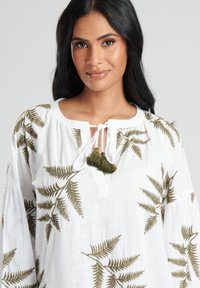 Vit linnetopp med grön ormbunke-broderi, nyckelhålsknäppning med vita band och små olivgröna tofsar vid öppningen.