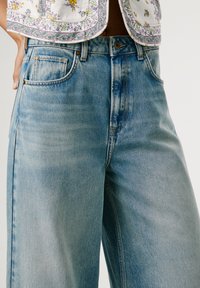 Svijetloplave traper hlače širokih nogavica s izblijedjelim završnim obradama, pet džepova i kopčanjem na gumb. Sadrže suptilne detalje šivanja uz šavove.