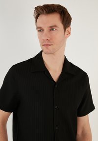 Camisa negra de manga corta con cuello tipo polo, que presenta una textura acanalada vertical y botones en la parte delantera. Tejido suave con un ligero brillo.