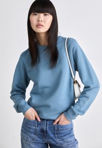 Sudadera de color azul claro con cuello redondo y puños acanalados, que presenta un logo en relieve en la parte delantera. Combinada con jeans de mezclilla de tiro alto.