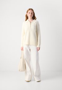 Pullover lavorato a maglia crema con colletto, chiusura a mezza zip e motivo testurizzato, abbinato a pantaloni bianchi morbidi e una borsa coordinata.