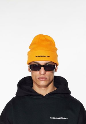 Orange hue strikhue med en tekstureret strik og sort logo, parret med sorte rektangulære solbriller og en sort hættetrøje.